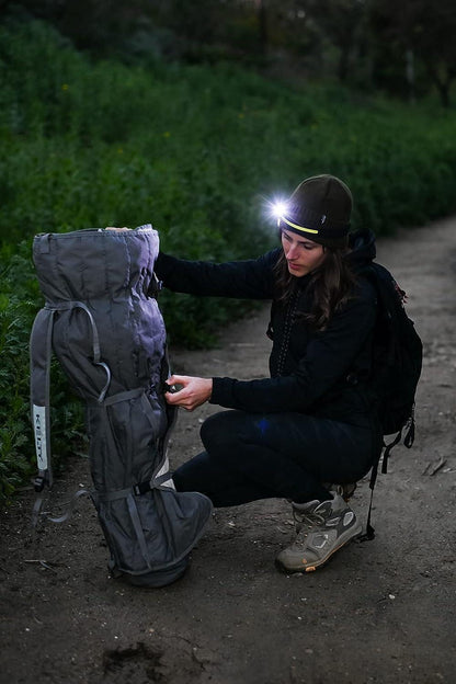 Person with a headlamp inspecting a gray bag outdoors on a path.